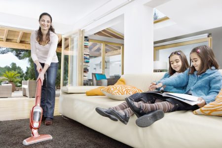 Mom cleaning the living room carpet with a stick vacuum while kids read on the sofa