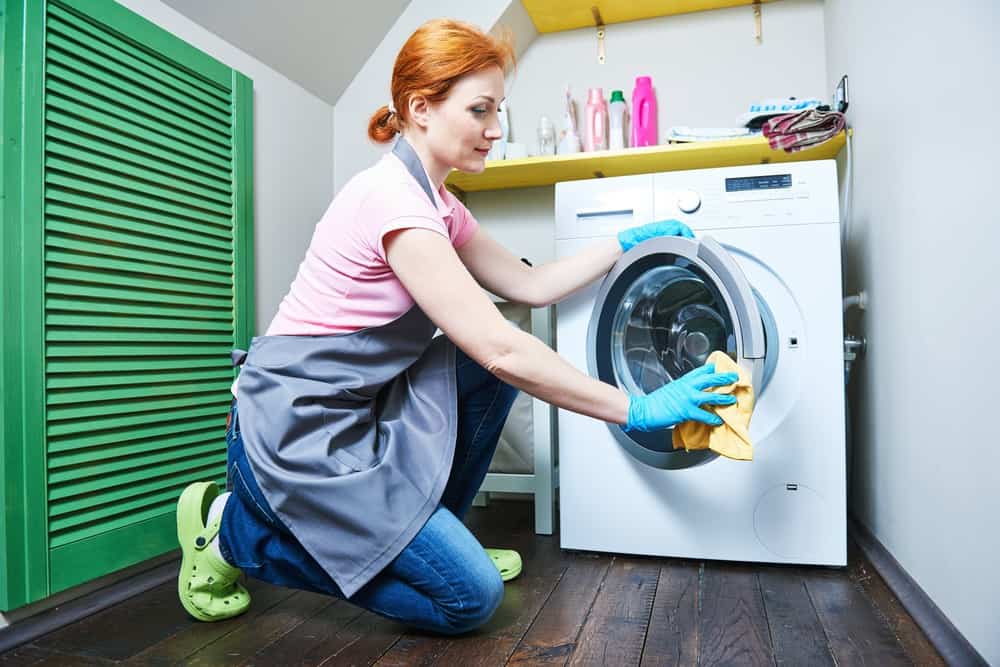 Woman in apron and gloves wiping washer with cloth in laundry room