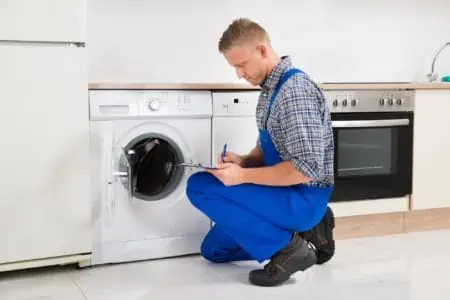 Man sitting in front of a washer while writing on clipboard with pen in the kitchen