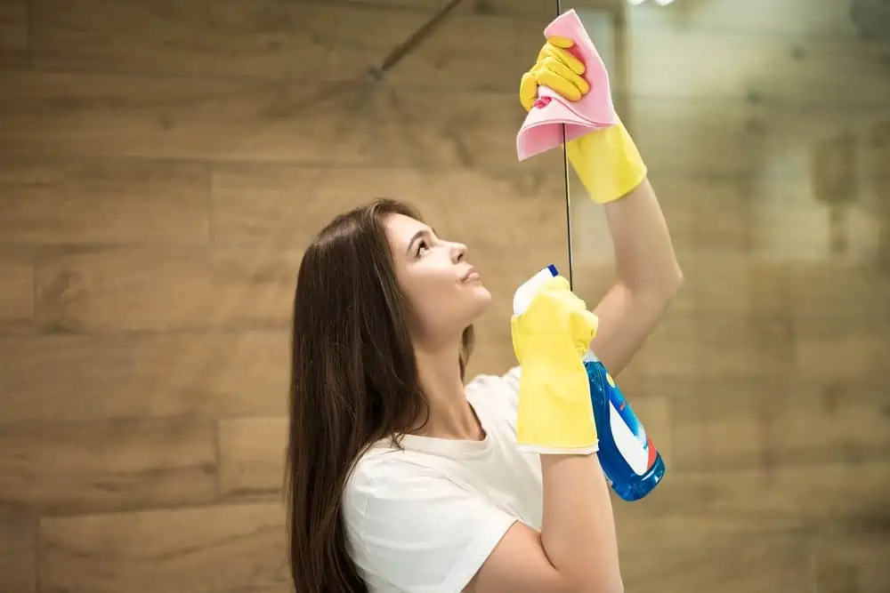 Woman in yellow gloves spraying detergent spray while wiping dust off from the shower door