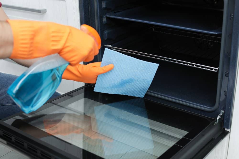 Man in orange gloves cleaning oven door with detergent spray and wipes