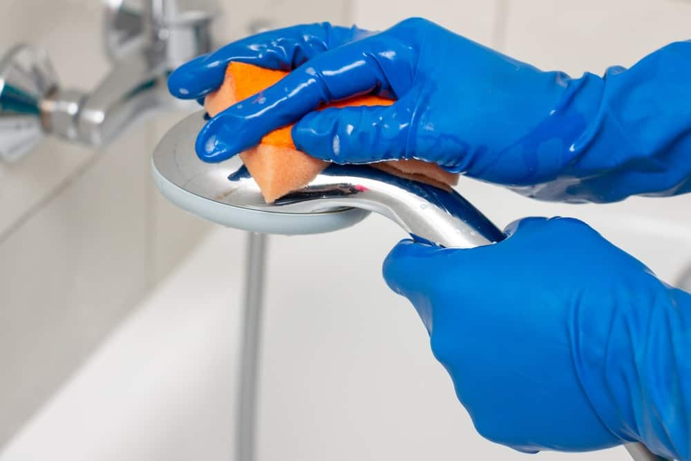 Hands wearing blue gloves cleaning shower head with orange sponge