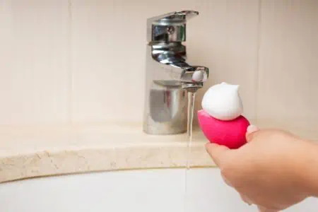 Woman's hand cleansing pink beauty blender with foam on top in the sink