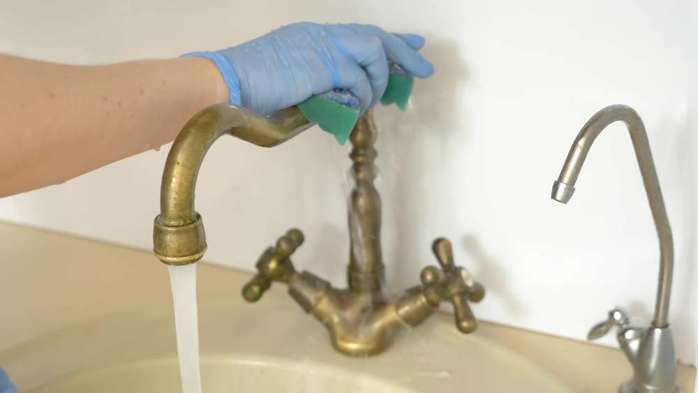 Female hands wearing rubber gloves cleaning brass faucet with green sponge