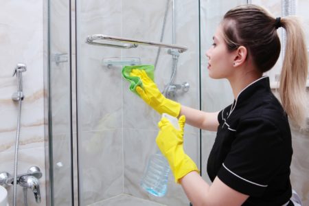 Woman in yellow gloves wiping glass shower door with green cloth and spray