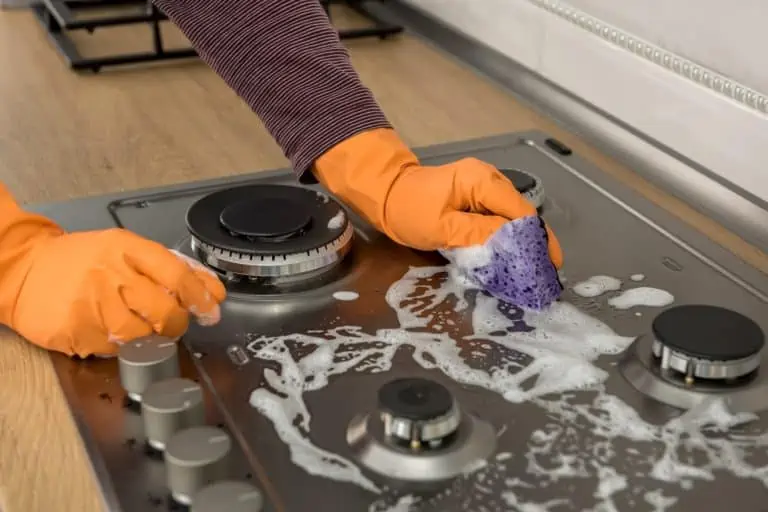 Female hand in orange gloves cleaning stainless steel gas stove with soap and sponge