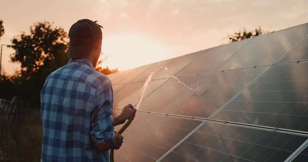 Male owner wearing cap washes home solar panels using water hose