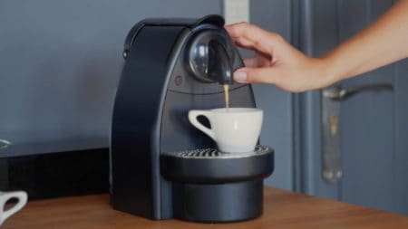 Woman's hand pressing button of nespresso machine with cup of coffee on top of wooden table