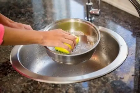 Woman washing aluminum pot in kitchen sink using soapy water and sponge