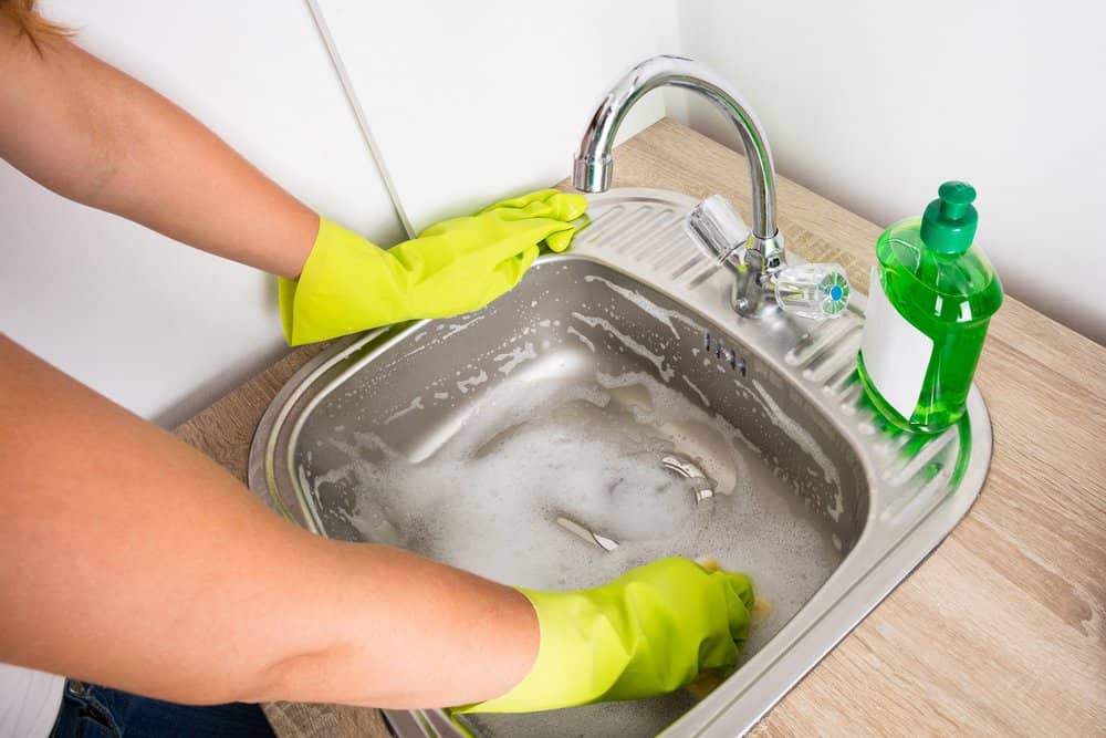 Woman's hand in gloves cleaning the stainless steel sink with sponge and detergent
