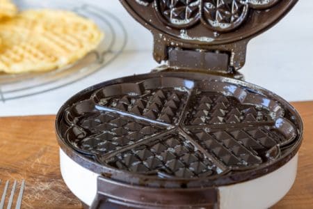 Empty dirty waffle maker on wooden table with waffle and fork in the background