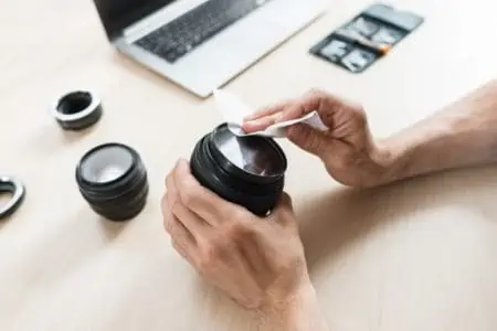 Male hands cleaning camera lens using white clean cloth on top table with laptop