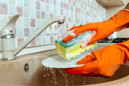 Woman hands in orange gloves washing dishes in the kitchen sink using sponge