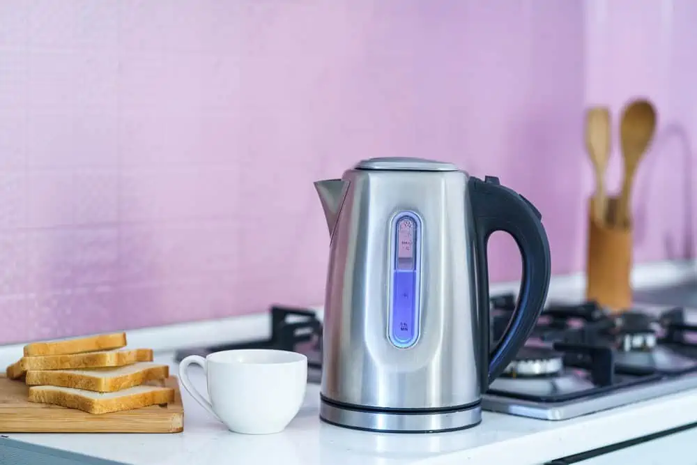 Electric kettle on kitchen countertop near the gas range with white cup and bread on board