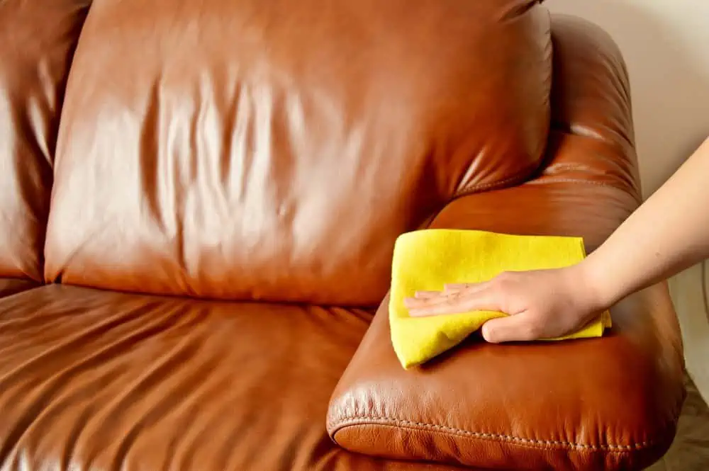 Female hand cleaning brown leather sofa with yellow microfiber cloth