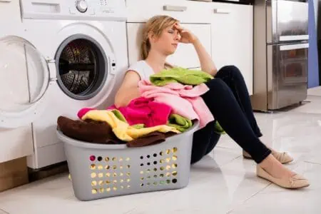 Frustrated woman sitting near washing machine with basket of dirty towels on the floor